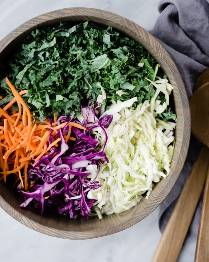 Thinly sliced kale, red cabbage, green cabbage, and shredded carrots in a wood serving bowl for kale slaw.