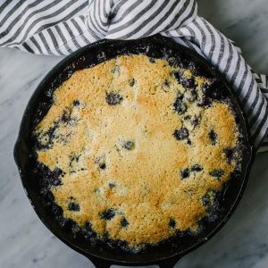 Gluten free blueberry cobbler cooked in a cast iron skillet on a marble table with a striped kitchen towel.