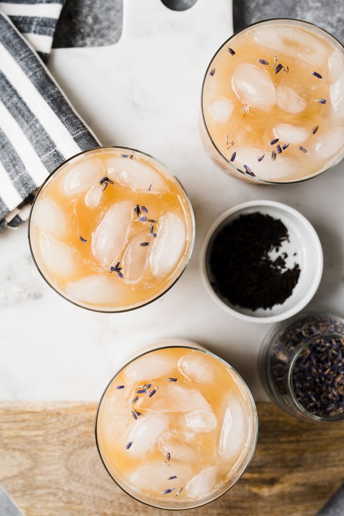 Three iced london fog tea lattes on a marble cutting board with a striped napkin and small jars of dried lavender and earl grey tea leaves.