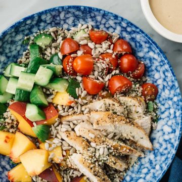 Crunchy summer quinoa salad in a blue bowl with chicken, peaches, cucumbers, tomatoes, sunflower seeds, hemp seeds, and a creamy sunbutter dressing.