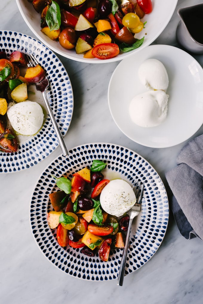 Two plates of stone fruit salad with tomatoes and burrata cheese on a marble table.