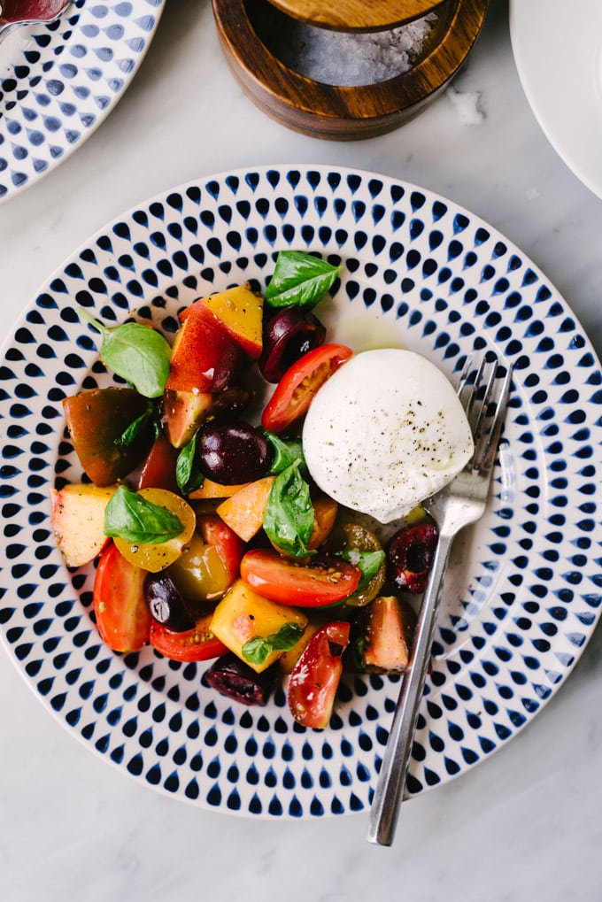 A serving of stone fruit salad with tomatoes and burrata cheese on a blue and white plate.