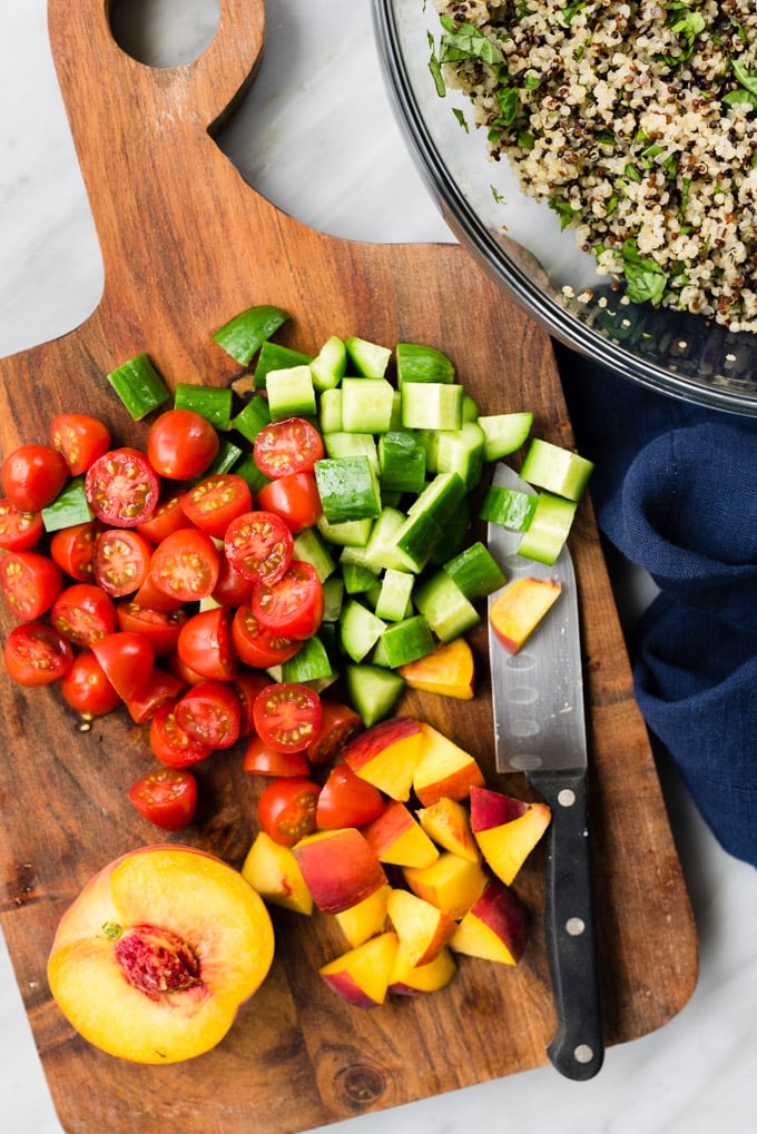 Diced peaches, cucumbers, and tomatoes on a wood cutting board next to a prep bowl of herb quinoa salad.