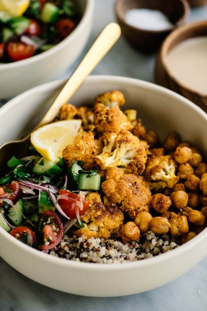 Roasted cauliflower shawarma with roasted crispy chickpeas, tomato salad, and quinoa in a cream bowl on a marble table.