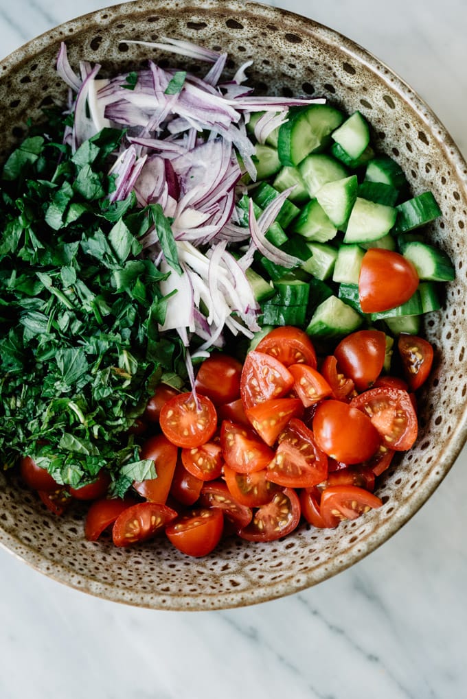Cucumbers, tomatoes, sliced red onion, mint, and parsley in a brown ceramic bowl for a vegan shawarma salad.