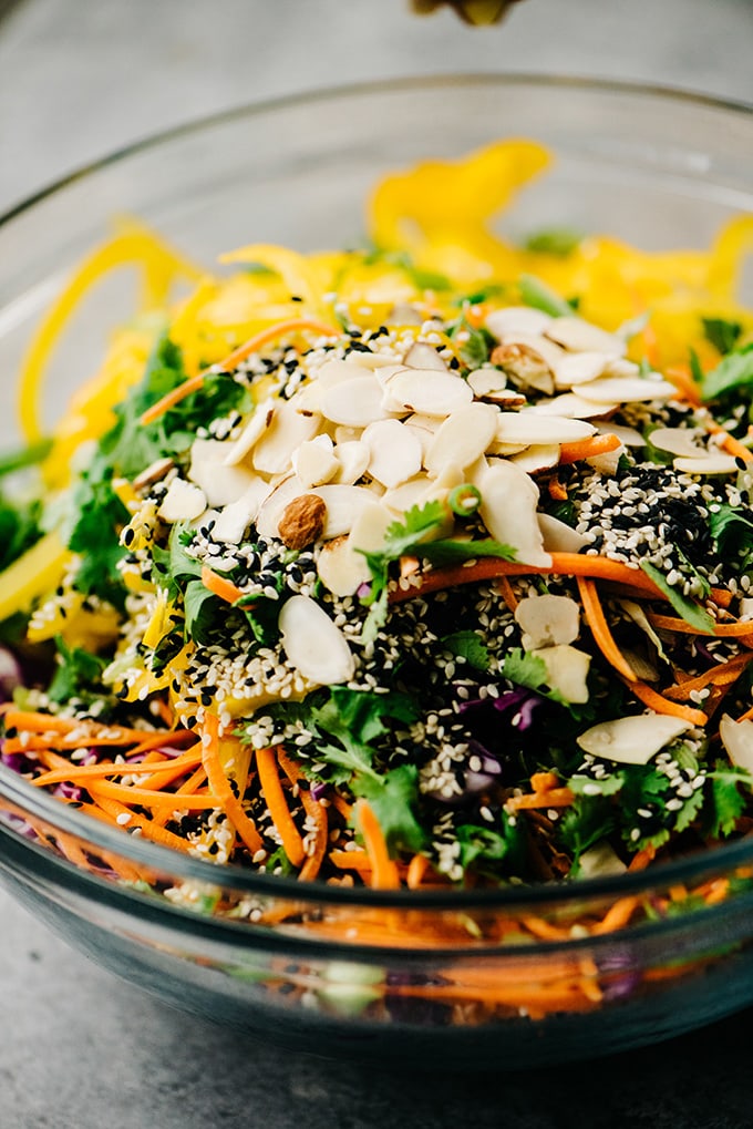 A glass mixing bowl filled with sliced vegetables, sesame seeds, and sliced almonds for a no mayo healthy chicken salad recipe.