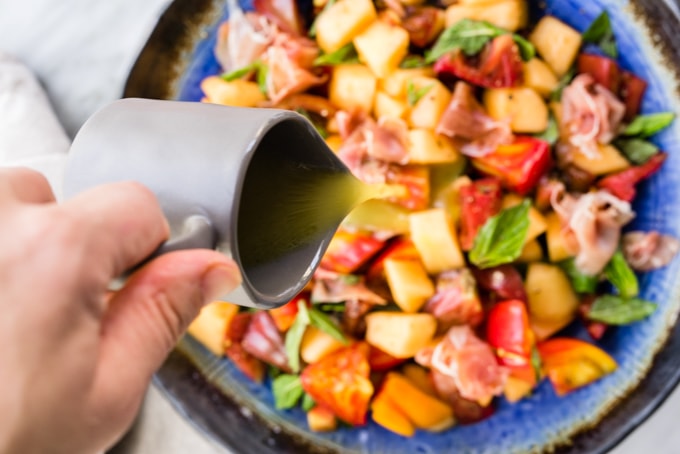 A woman pouring apple cider vinaigrette over a melon salad tossed with prosciutto, tomato, mint, and basil.