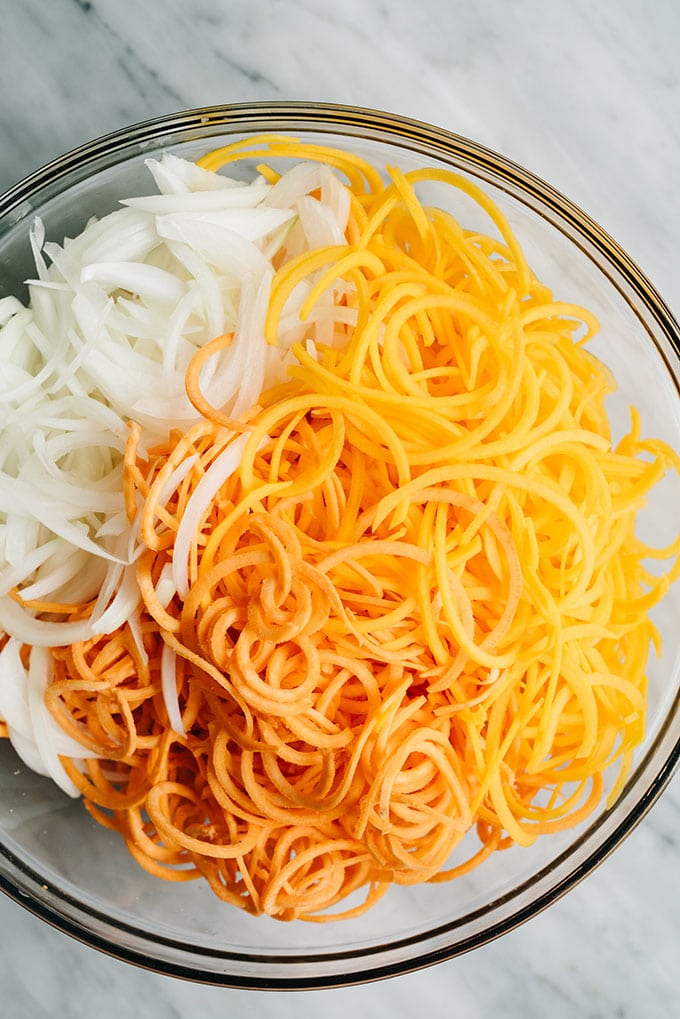 Spiralized sweet potatoes and butternut squash with thinly sliced sweet onion in a glass mixing bowl.