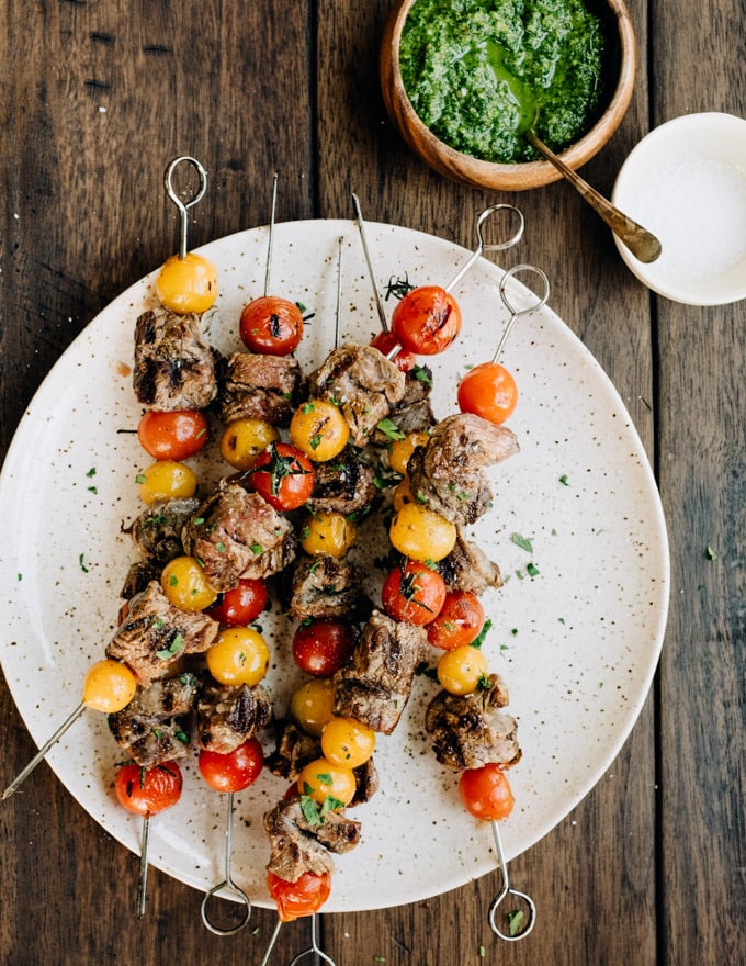 A platter of grilled steak skewers with tomatoes and a side of dairy free parsley pesto.