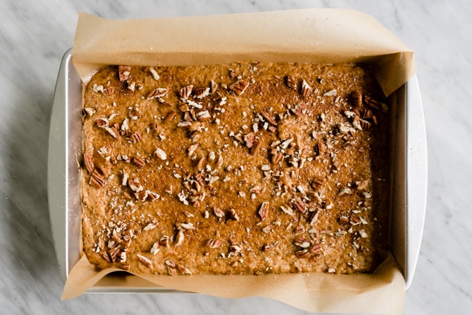 A cake pan lined with parchment paper, filled with healthy pumpkin bar batter, topped with chopped pecans.