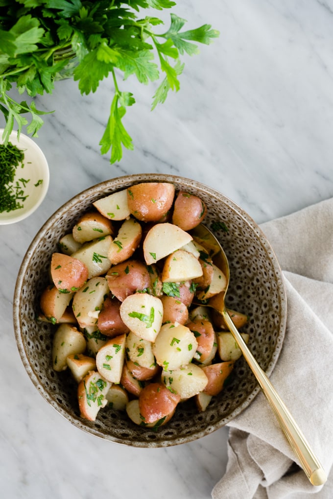 A brown serving bowl filled with italian potato salad on a marble table with a bundle of fresh parsley.