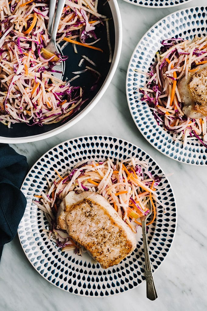 Two plates of thick cut oven roasted pork chops with apple jicama slaw on a marble table.
