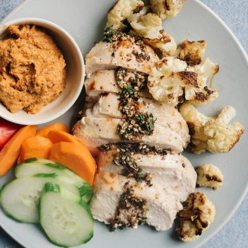 A plate of baked zaatar chicken with roasted cauliflower, muhammara dipping sauce, and sliced raw vegetables.
