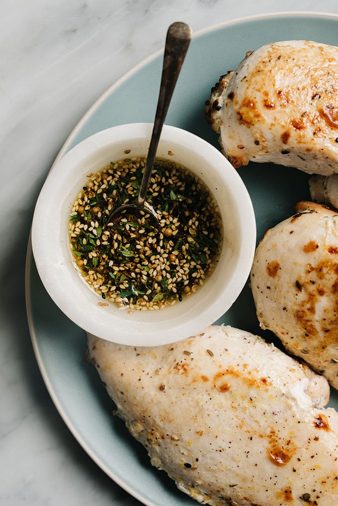A small marble bowl of zaatar spice mixed with olive oil on a platter with baked zaatar chicken breasts.