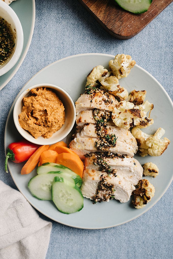 A plate of baked zaatar chicken with roasted cauliflower, muhammara dipping sauce, and sliced raw vegetables.