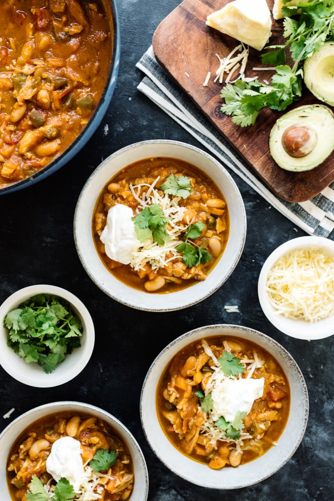 Several bowls of pumpkin chicken chili surrounded by toppings and garnishes such as avocado, sour cream, sharp cheddar and cilantro.