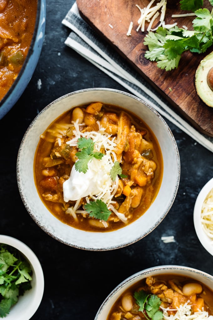 A bowl of gluten free pumpkin chicken chili on a black surface surrounded by small bowls of toppings. 