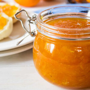 A jar of homemade orange marmalade on a white background with orange slices in the background.