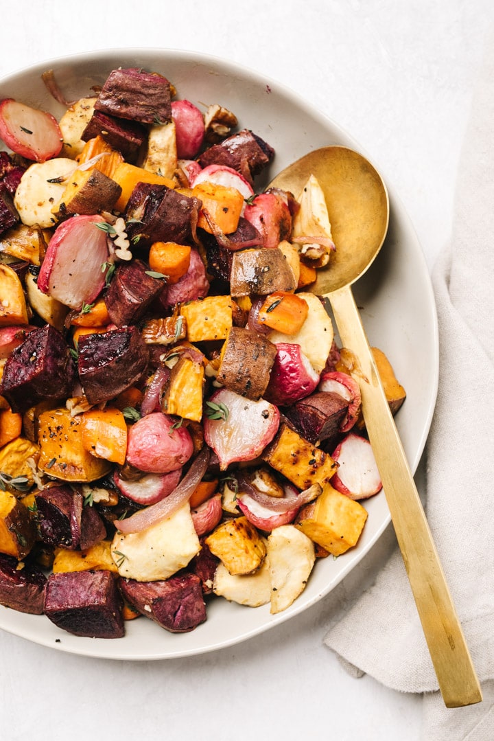 A tan serving bowl filled with maple roasted root vegetables with a gold serving spoon and linen napkin on a concrete background.