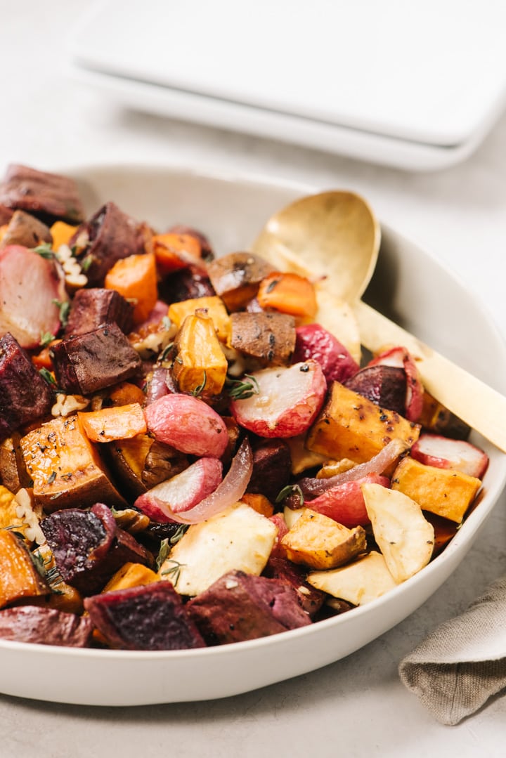 Side view, roasted root vegetables with maple syrup in a tan serving bowl with small square plates stacked in the background.