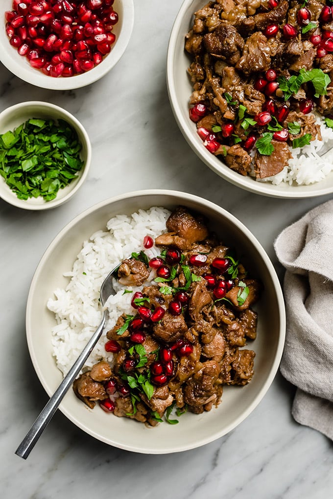 Two bowls of pomegranate chicken over white rice on a marble table with small bowls of pomegranate seeds and fresh parsley, along with a linen napkin.