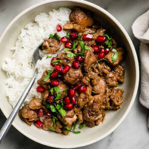 Pomegranate chicken over white rice in a low tan bowl, garnished with pomegranate seeds and fresh parsley.