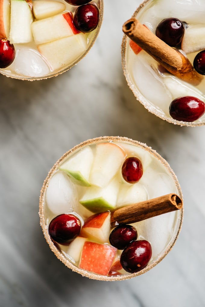 An overhead view of three apple cider sangria cocktails on a marble table with diced apples, whole cranberries, and a cinnamon stick garnish.