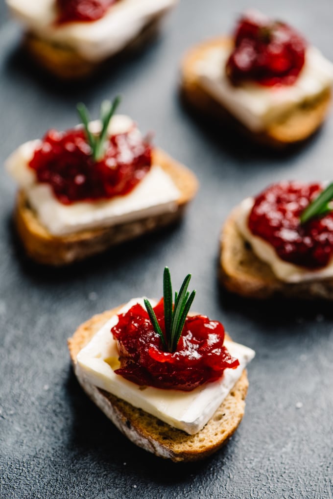 Cranberry brie bites cocktail appetizer spread - toasted baguettes topped with a slice of brie, cranberry jam, and a sprig of rosemary garnish.