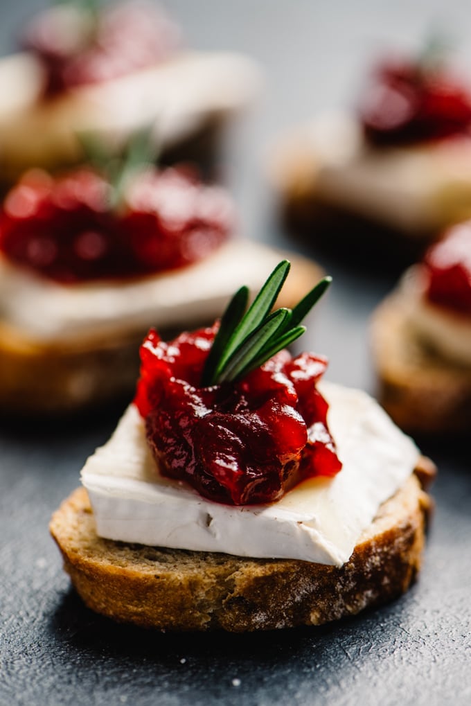 A cranberry brie bite - toasted baguette topped with a slice of brie cheese, cranberry jam, and rosemary garnish.