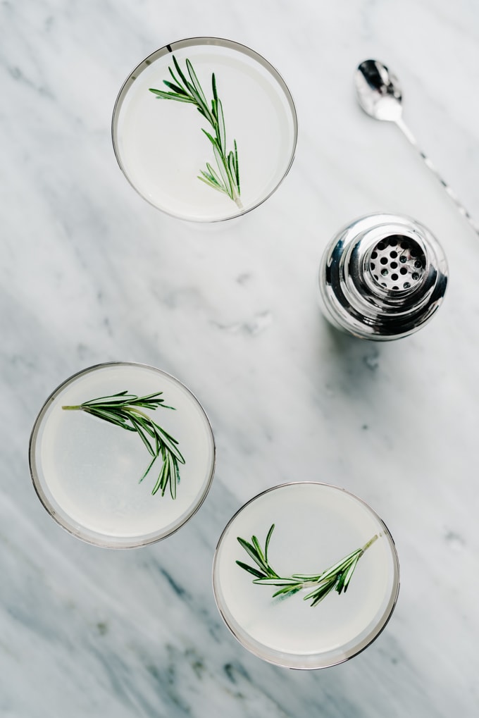 An overhead view of three rosemary vodka gimlet cocktails on a marble table with a silver cocktail shaker.