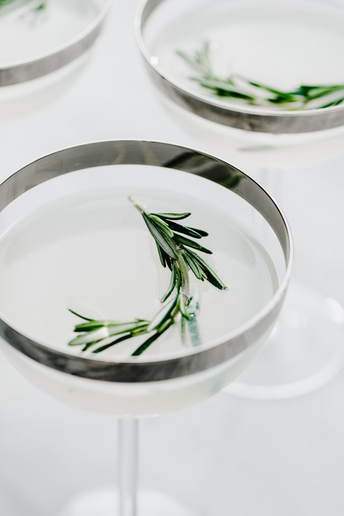 Three coupe glasses with silver rims filled with a straight up rosemary vodka gimlet garnished with rosemary sprigs.