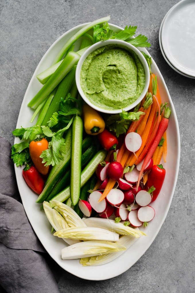A large white platter of sliced vegetable crudite with a bowl of white bean green goddess dip and several small side plates.