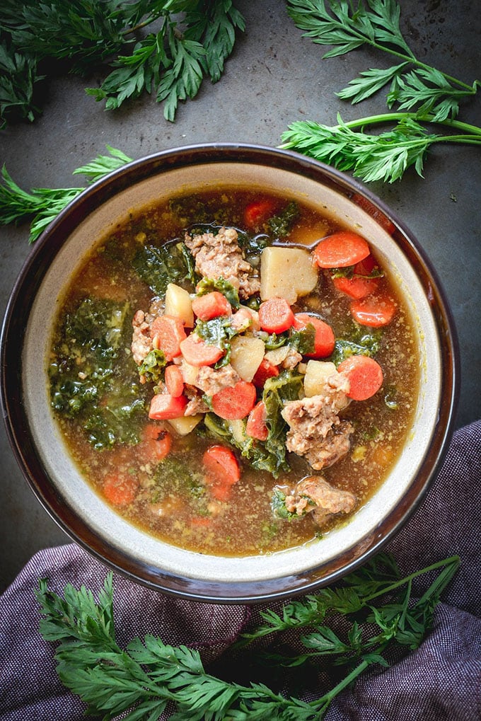 Sausage, kale, and carrot whole30 soup in a brown iand white bowl with a side of fresh herbs.