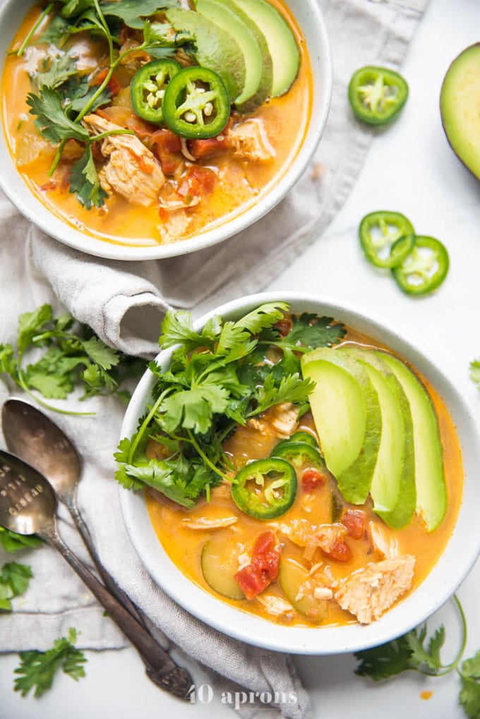 Two bowls of chicken tortilla less soup with avocado, cilantro and jalapeno on a marble table.