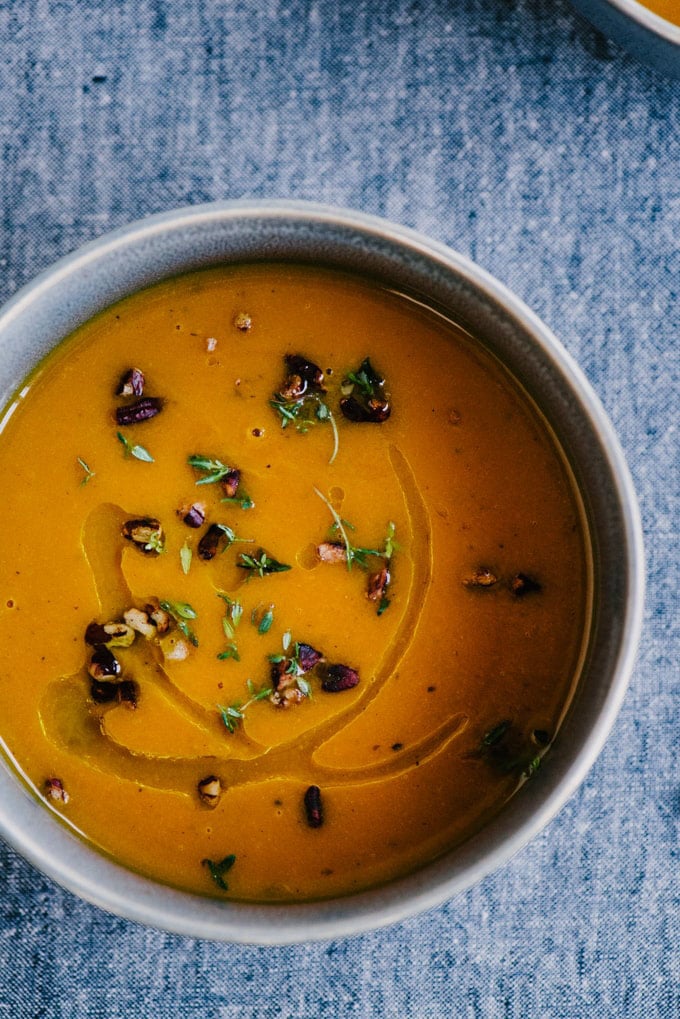 A bowl of carrot and apple whole30 soup garnished with pecans and thyme on a blue tablecloth. 