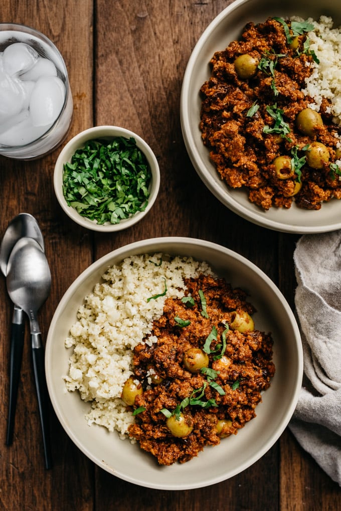 Two bowls of Whole30 picadillo over cauliflower rice with fresh cilantro on a wood table.
