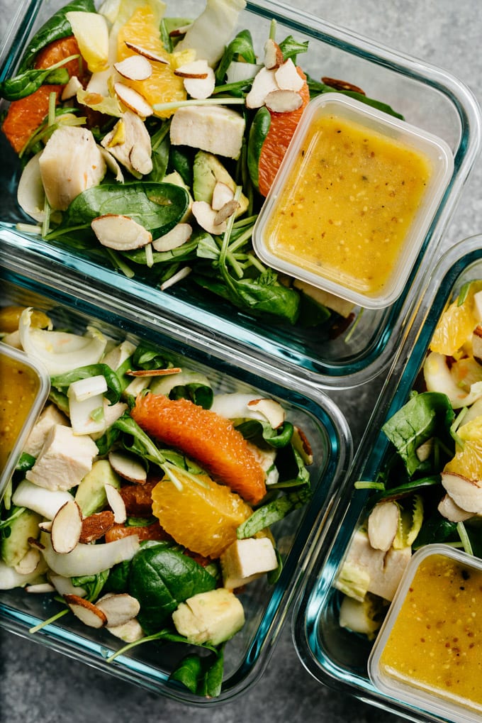 Three meal prep containers filled with orange chicken salad and a small container of citrus vinaigrette.