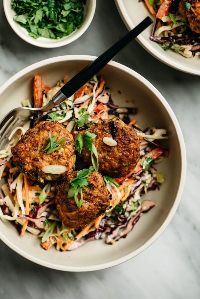 A bowl of Whole30 ginger pork poppers with citrus asian slaw on a marble table with small bowls of cilantro and green onion.
