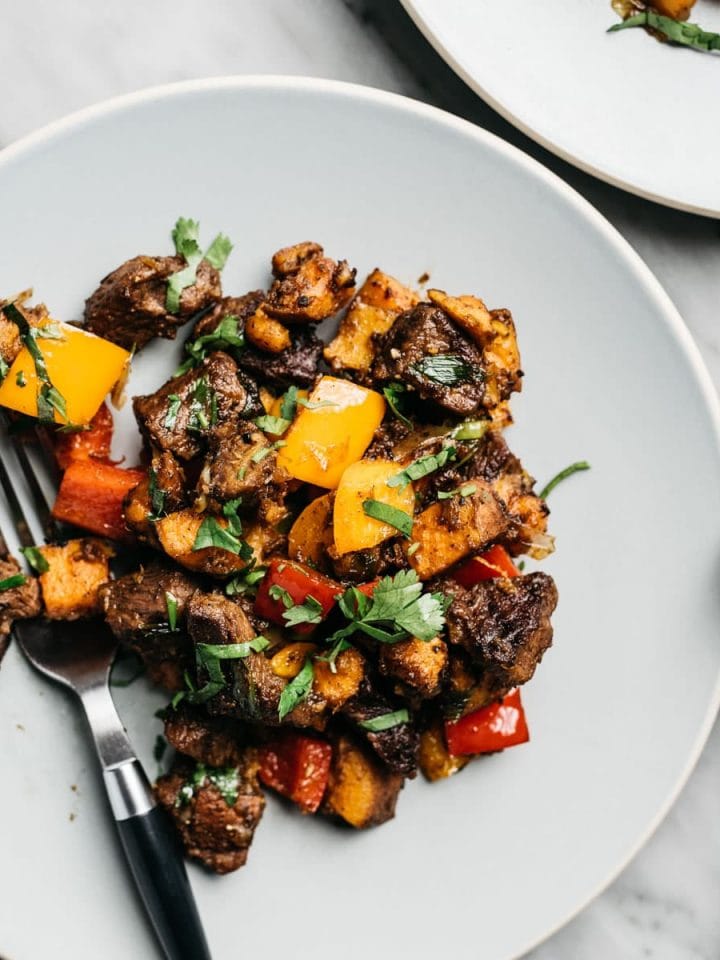 Whole30 steak bites sauteed with sweet potatoes and bell peppers on a blue plate with a black fork.