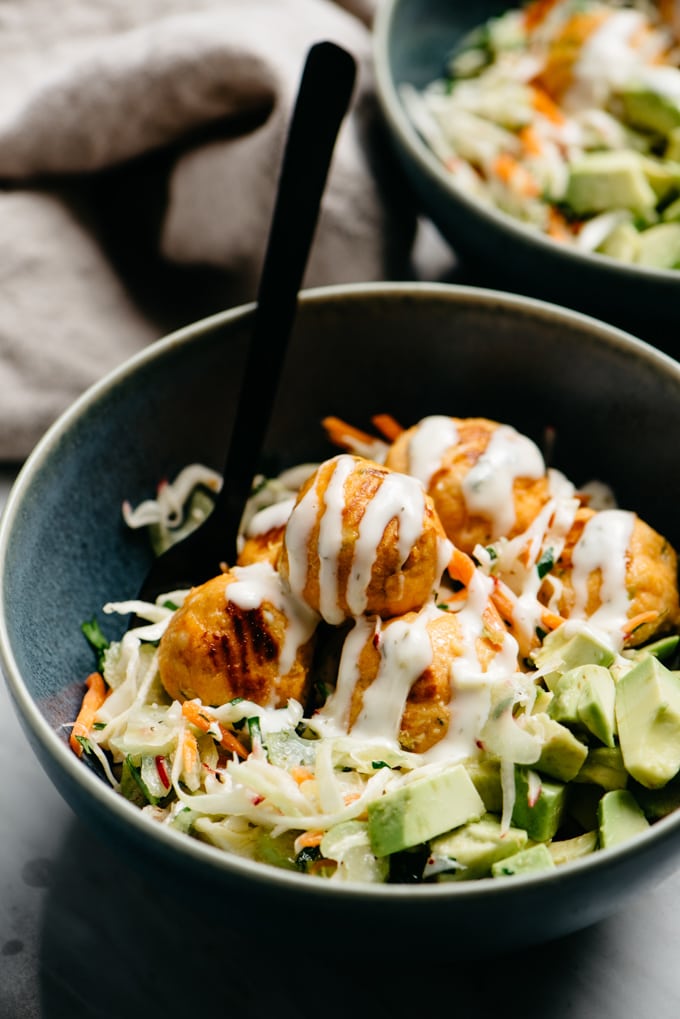 Gluten free buffalo chicken meatballs over a bed of no mayo coleslaw with avocado and ranch dressing in a blue bowl.