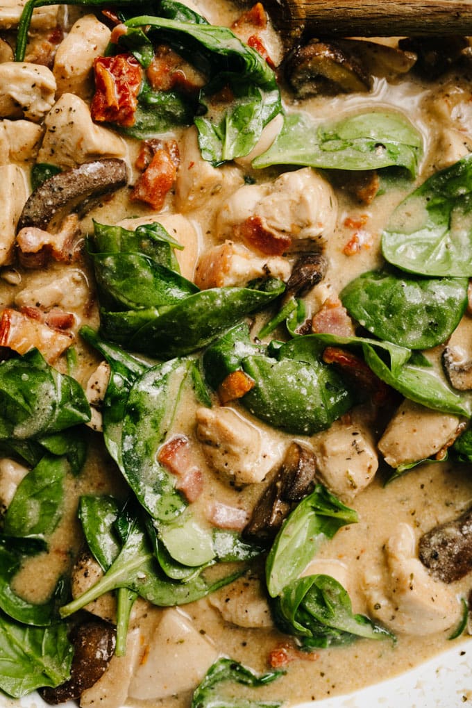 Healthy tuscan chicken simmering in a skillet with sun dried tomatoes, mushrooms, spinach, and a creamy dairy free bacon sauce.