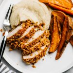 Baked pecan crusted chicken over cauliflower puree with baked sweet potato fries on a white plate.