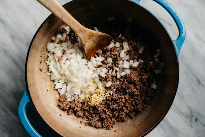 Ground beef in a blue dutch oven with raw onions, ginger, and garlic for preparing keema curry.