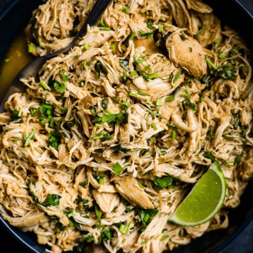 A serving spoon tucked into a bowl of shredded slow cooker cilantro lime chicken, garnished with a lime wedge and lots of fresh cilantro.