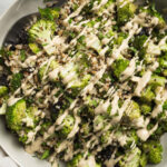 A serving spoon tucked into a large tan serving bowl filled with broccoli quinoa salad - roasted broccoli, quinoa, sunflower seeds, dried cherries, mint, and creamy sunbutter dressing.