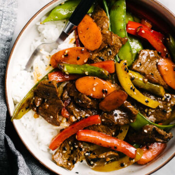 Steak stir fry with rainbow vegetables over steamed white rice in a low bowl, garnished with sesame seeds.