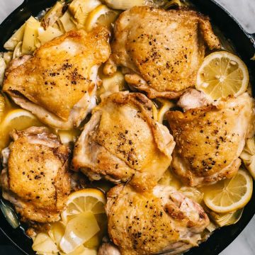Braised artichoke chicken with lemons, garlic, and tarragon in a skillet on a marble table with a side of roasted potatoes.