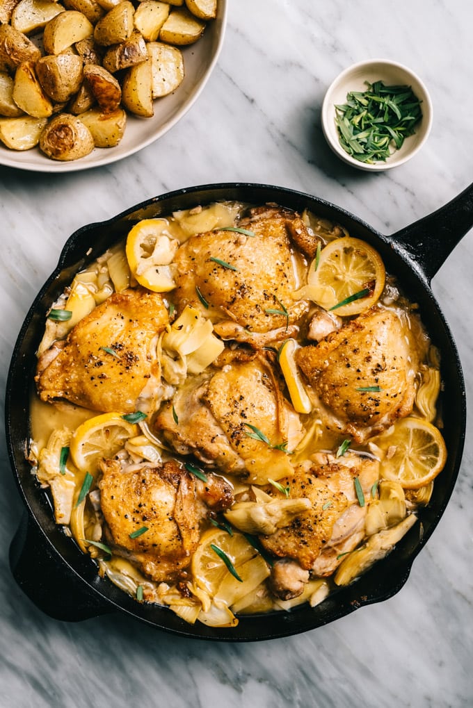 One-pan artichoke chicken with lemons, garlic, and tarragon in a skillet on a marble table with a side of roasted potatoes.