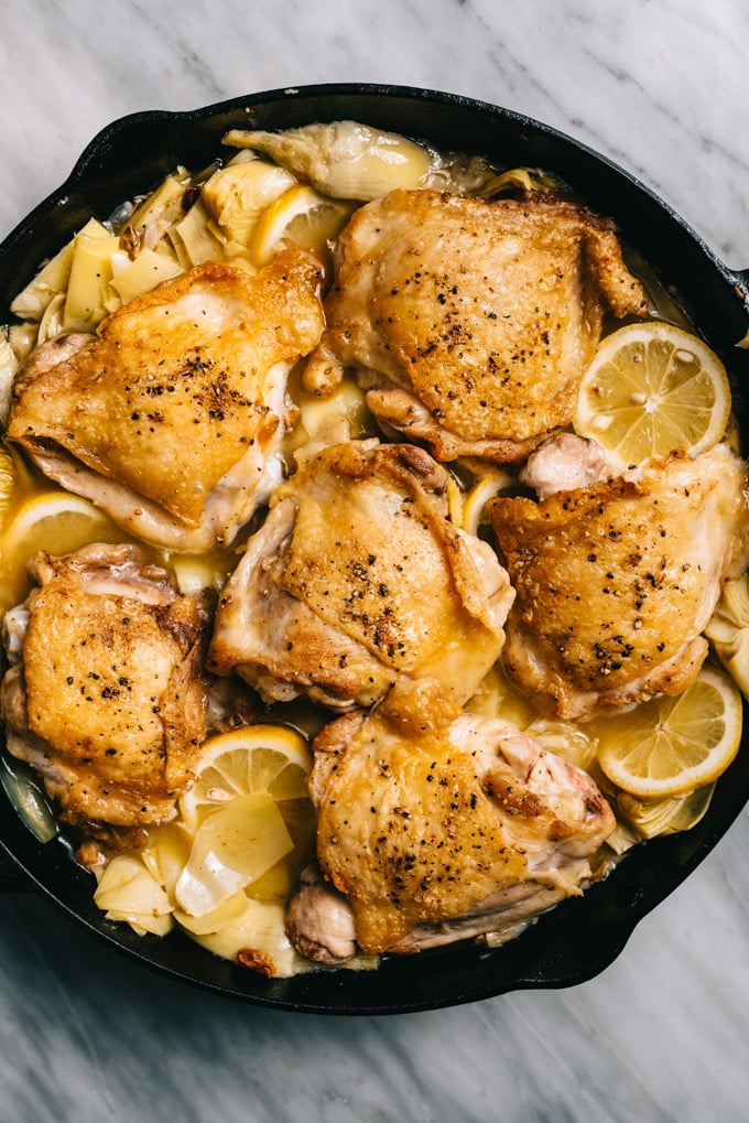 Chicken thighs in a skillet with artichoke hearts, lemon slices, garlic, and white wine sauce.