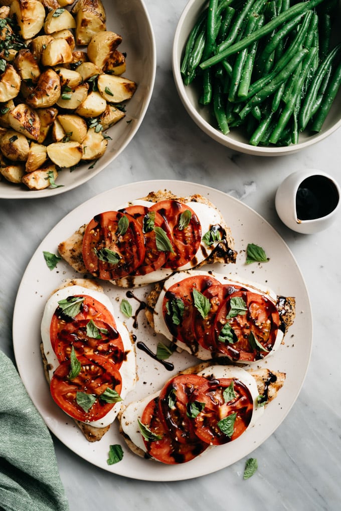 A platter of caprese chicken with tomato, basil, and mozzarella on a marble table with side bowls of roasted potatoes and green beans. 
